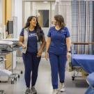 Two healthcare workers in navy scrubs walking and smiling down a hospital corridor